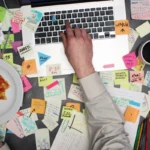 Top view on multitasking man working on laptop computer with post it notes all around his office desk.