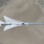 NASA’s white, blue, and red X-59 research aircraft flies over the Mojave Desert near NASA’s Armstrong Flight Research Center in Edwards, California, during its third flight. The aircraft is centered in the frame, viewed from above with the canopy, both wings, NASA and X-59 logos visible, and the landing gear down with one wheel visible. Below, rugged terrain transitions into the smooth texture of a dry lakebed as the aircraft, nearly 100 feet long, flies toward the right side of the frame.
