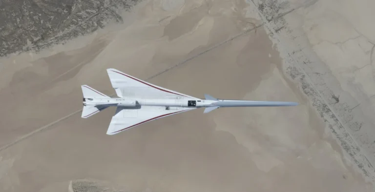 NASA’s white, blue, and red X-59 research aircraft flies over the Mojave Desert near NASA’s Armstrong Flight Research Center in Edwards, California, during its third flight. The aircraft is centered in the frame, viewed from above with the canopy, both wings, NASA and X-59 logos visible, and the landing gear down with one wheel visible. Below, rugged terrain transitions into the smooth texture of a dry lakebed as the aircraft, nearly 100 feet long, flies toward the right side of the frame.
